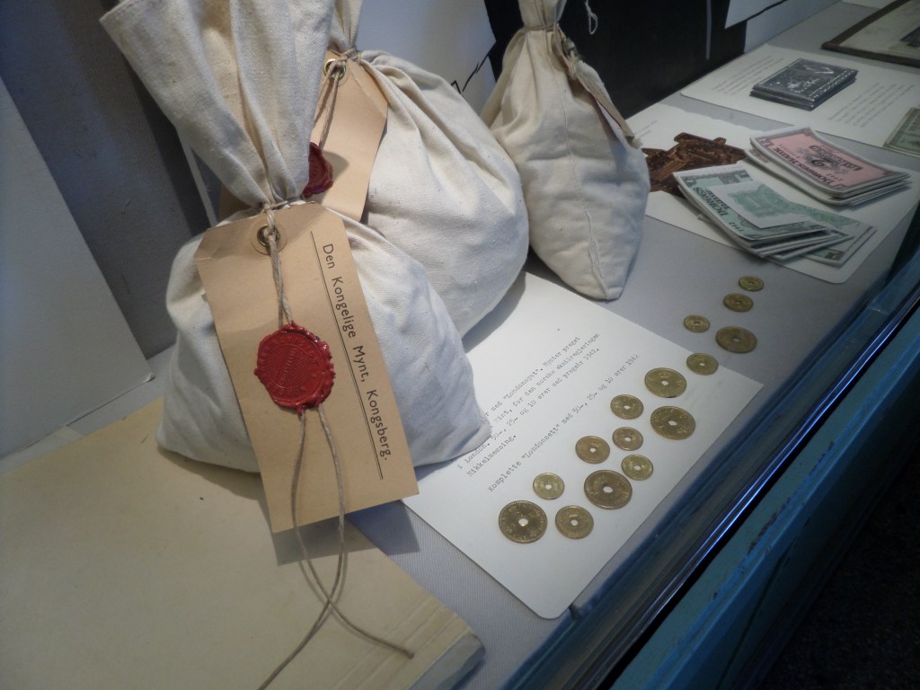 Kuvassa kankaisia pusseja ja kultakolikoita, joiden keskellä on pyöreä reikä. Pusseissa on pahvilappu ja sinetti, joka kertoo rahojen kuuluvan kuninkaalle.