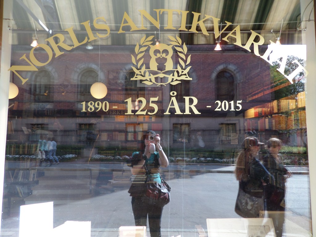 Kuva Norlis Antikvariaatin lasi-ikkunasta, johon heijastuu myös kolme ihmistä - yksi näistä on kuvaa ottava Katja.