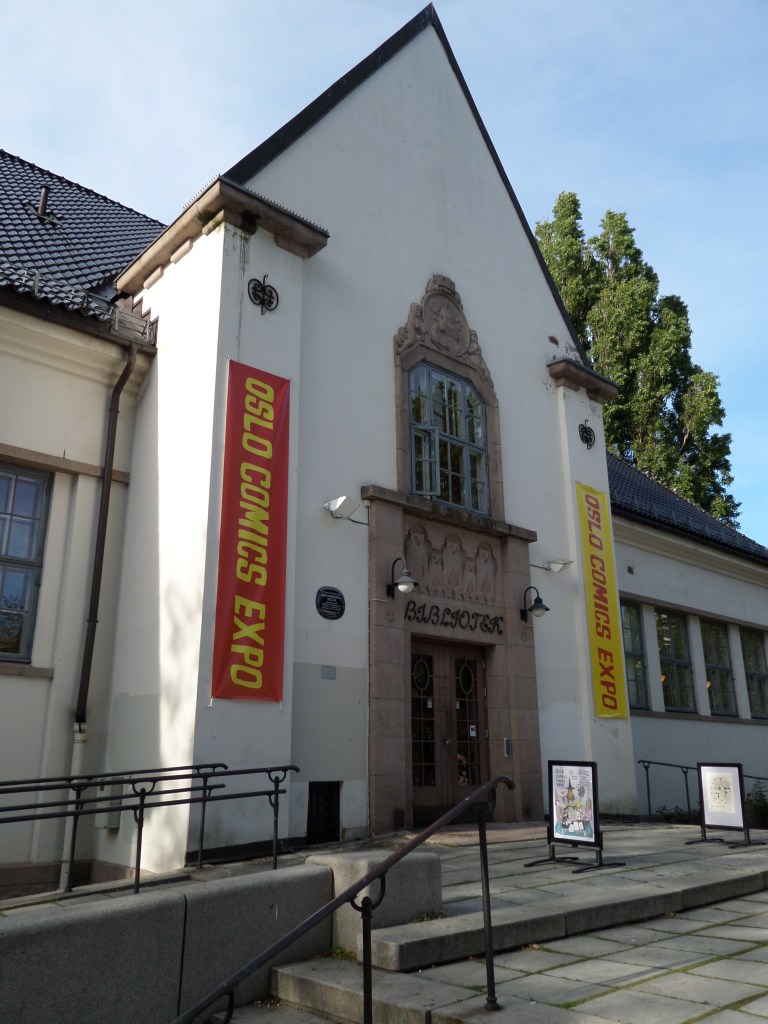 Kuva valkoisesta talosta, jona isossa, koristeellisessa oviaukossa lukee BIBLIOTEK. Oviaukon kummallakin puolella on mainoslakanat, joissa lukee OSLO COMICS EXPO.