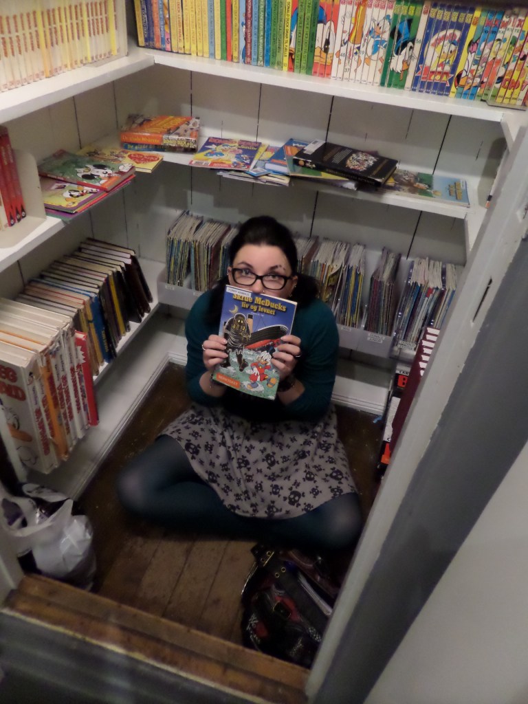 Kuva Aku Ankka-sarjakuvia täynnä olevasta pienestö komerosta, jossa on valkoiset hyllyt. Puisella lattialla istuu Katja, joka on puoliksi piiloutunut Don Rosan piirtämän sarjakuvan taakse.