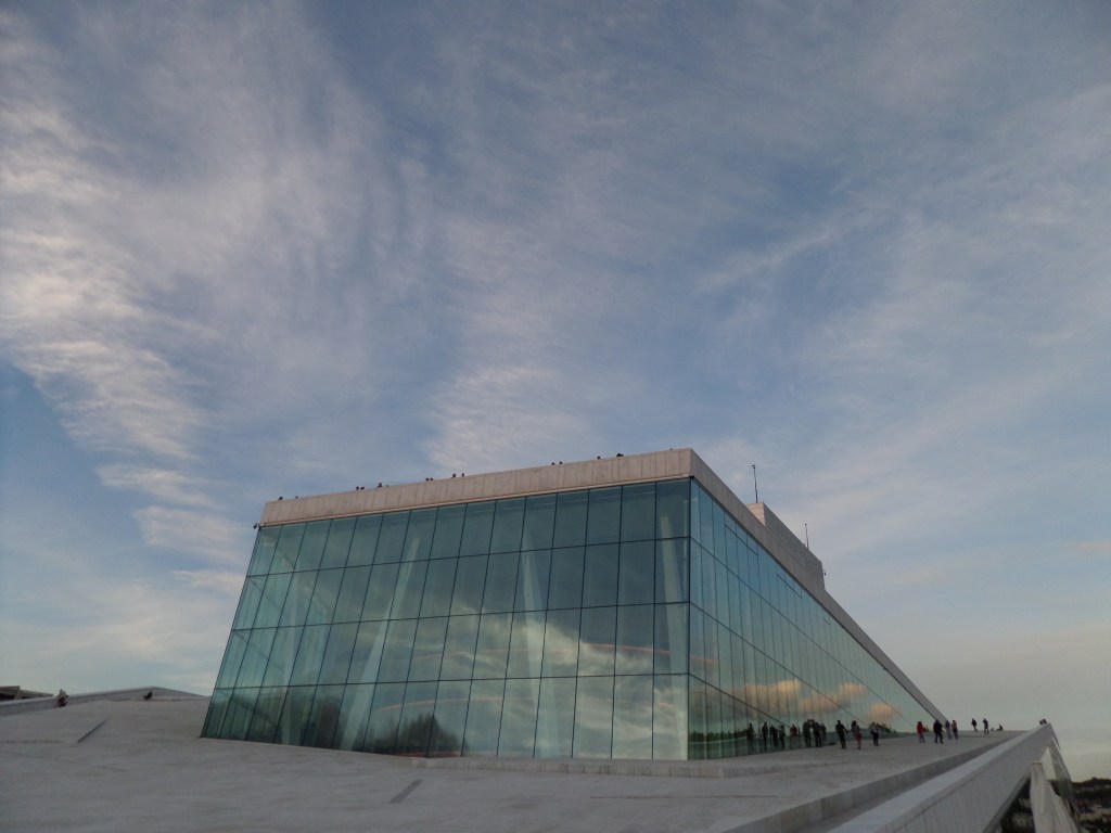 Kuva Oslon oopperatalon lasisesta seinästä ja sitä kiertävästä leveästä "rampista". Taivas on sininen ja täynnä utuisia pilviä. Joitakin ihmisiä on kävelemässä ramppia pitkin rakennuksen katolle.