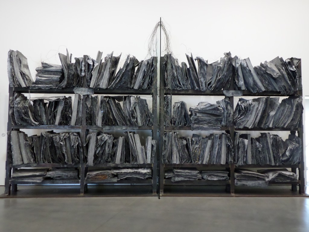 Kuva taideteoksesta, joka on kuin valtava kirjahylly täynnä todella isoja, paksuja kirjoja. Taideteos näyttää siltä, kuin se olisi palanut täysin ja jäljellä olisivat kärähtäneet teokset.