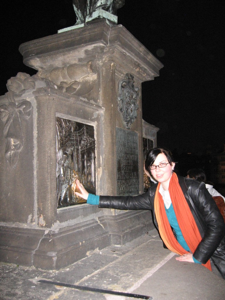 Illan pimeydessä otettu kuva Katjasta, joka ojentaa oikeaa kättään koskemaan kivestä tehtyä reliefiä Kaarlen sillalla. Katjalla on musta nahkatakki ja pitkä oranssi huivi.