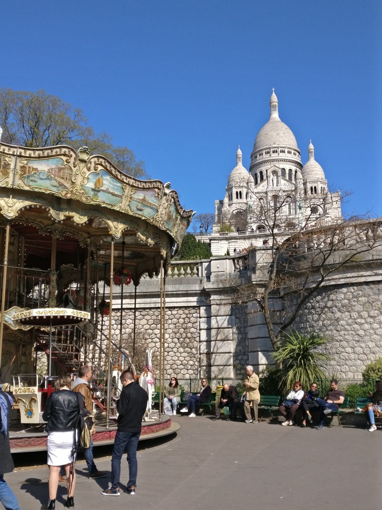 Kuvassa etuvasemmalla osittain näkyvä iso karuselli, jonka takana suurehko kivimuuri. Kivimuurin takana korkealla nousee korkeuksiin kirkko, jossa on kolme pyöreää kupolia. Kirkko on väriltään vaaleanharmaa ja se erottuu hyvin sinistä taivasta varten. Kuvassa on jonkin verran ihmisiä istumassa penkeillä ja seisomassa karusellin edessä.