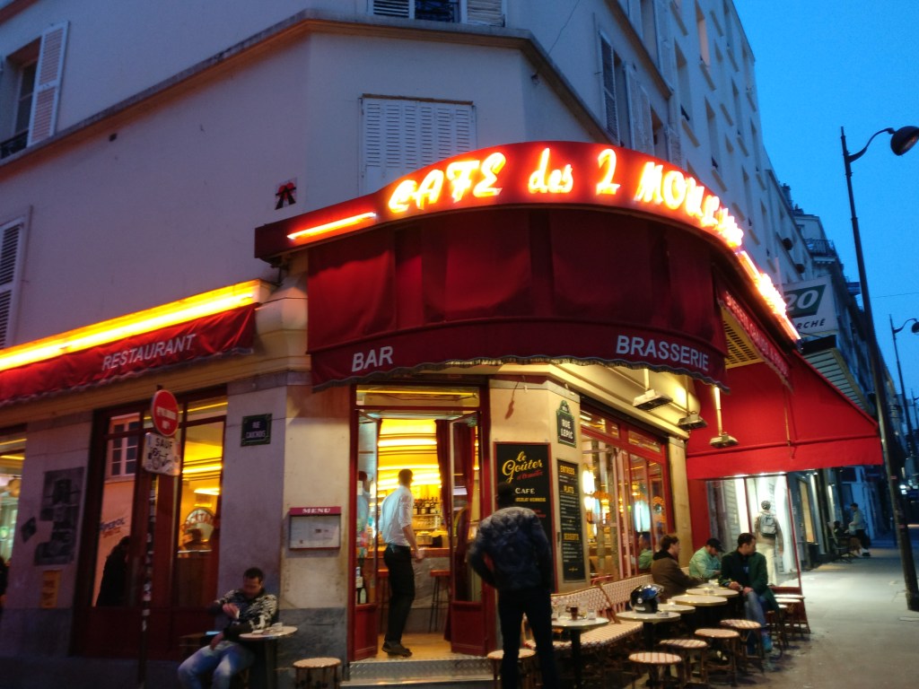 Kuva rakennuksen kulmasta, jossa on punaisella markiisilla koristeltu kahvila. Kahvilan nimikyltti loistaa markiisin huipulta. Ikkunoiden ulkopuolella on pieniä pöytiä ja tuoleja, joilla istuu ihmisiä.