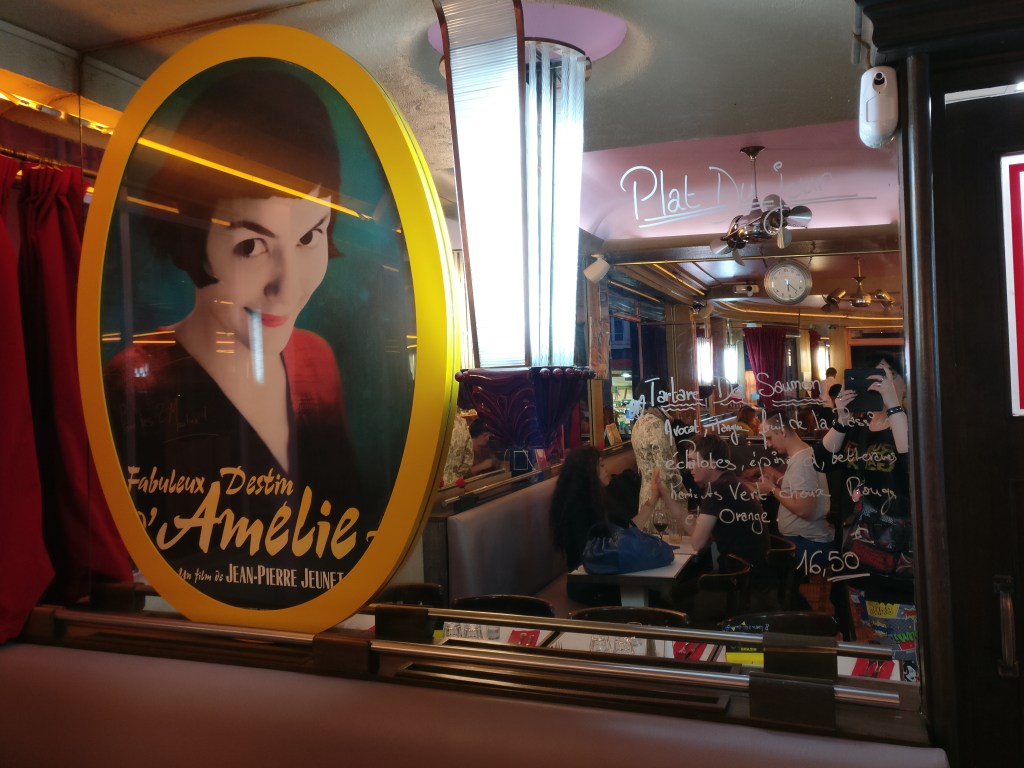 Kuva isosta peilistä, johon on kirjoitettu päivän ruoka-annokset ranskaksi. Tekstin vieressä on soikion mallinen kuva Amélie-elokuvan kansikuvasta, jossa Amélie hymyilee kameraan.