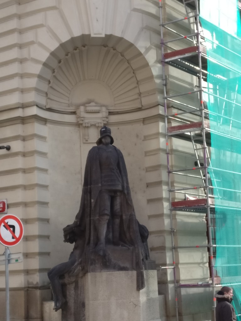 Kuva talon nurkalla seisovasta mustasta patsaasta, jolla on yllään haarniska, viitta ja kasvot peittävä kypärä.