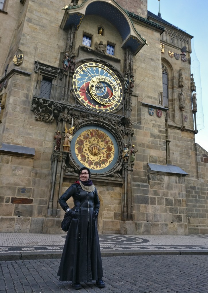 Kokovartalokuva Katjasta ison rakennuksen edessä. Katjalla on pitkä musta nahkatakki ja neulottu kaulaliina kaulassa. Taustalla näkyy astronominen kello, jossa on kaksi päällekkäistä kellotaulua: alemmassa on esillä horoskooppimerkit, ylemmässä aurinkokello.