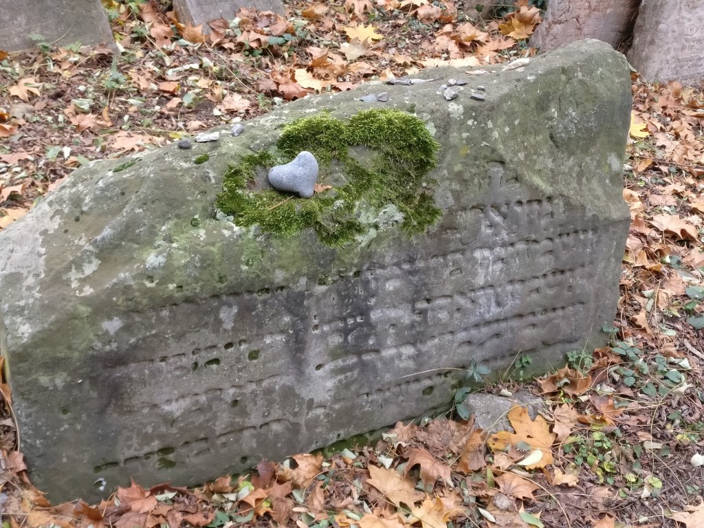 Kuva vanhasta hautakivestä, jossa on kulunutta tekstiä - kenties hepreaksi. Kiven päällä on vihreää sammalta, jonka keskelle on jätetty sydämenmuotoinen kivi.