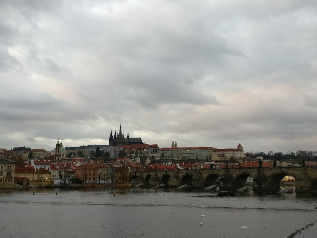 Kuvassa keskellä levittäytyy Kaarlen silta. Etualalla joki, tausalla Prahan linna, jonka keskellä seisoo Pyhän Vituksen katedraali. Sää on pilvinen ja synkähkö.