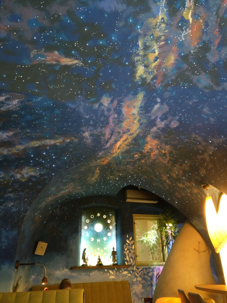 Kuva ravintolan katosta, joka on maalattu moniväriseksi muistuttamaan linnunrataa. Katto on hieman kupera ja takaseinällä näkyy valaistuja tauluja, buddha-patsauta sekä looseja ruokailijoille.