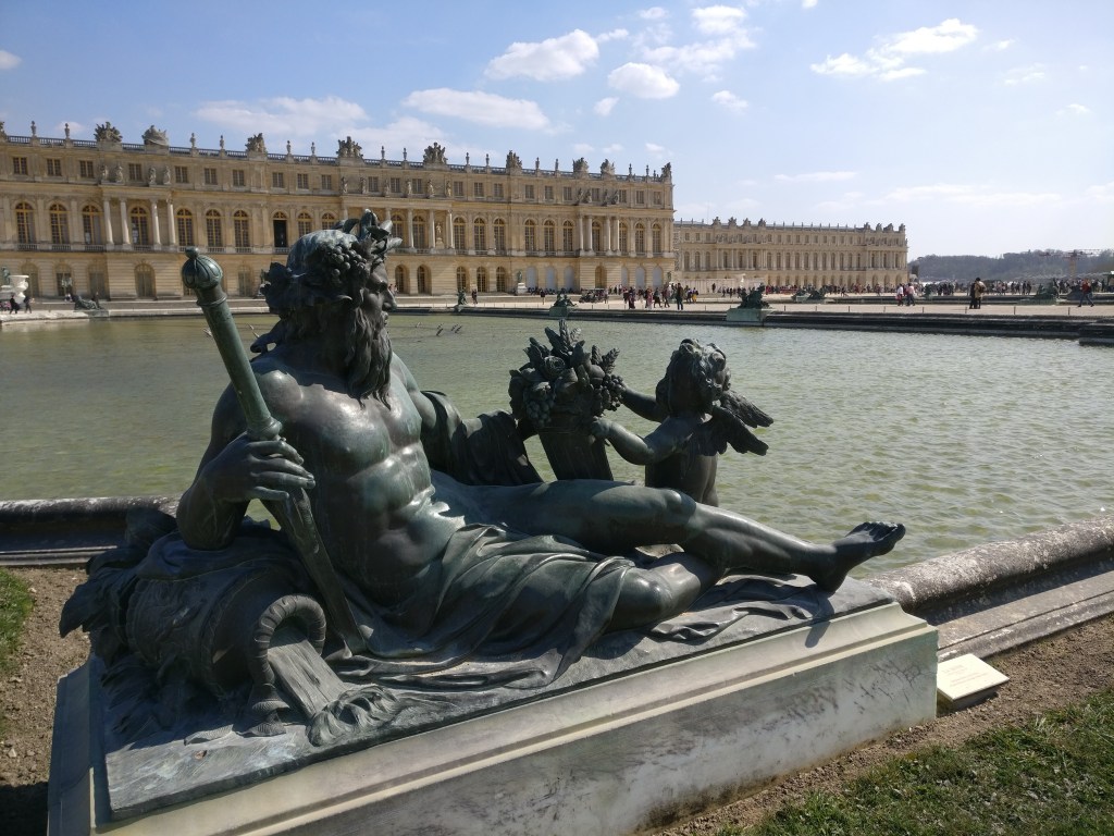 Kuva melkein kyljellään pötköttelevästä parrakkaasta patsaasta, joka pitelee kädessään jonkinlaista valtikkaa ja nojailee kaatuneeseen ruukkuun. Hahmon päässä on seppele ja hän on ottamassa vastaan jonkinlaista kimppua pieneltä enkelihahmolta. Patsas on ison vesialtaan reunalla. Taustalla on Versailles'n palatsi.