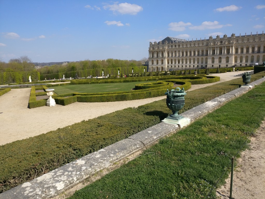 Vihreitä, kulmikkaisiin muotoihin leikattuja pensasaitoja, joiden joukossa on jonkinlaisia maljakkoja. Taustalla näkyy osa Versailles'n palatsia.