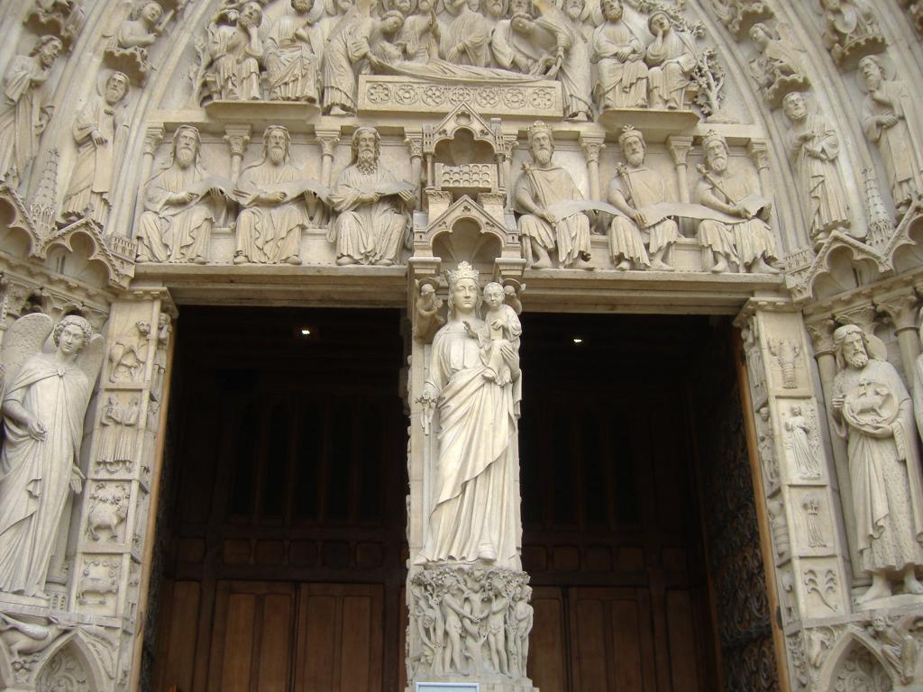 Valokuva Notre Damen pääovista, joiden keskellä on valkoinen Neitsyt Marian patsas. Marialla on kruunu päässään ja hän pitelee Jeesus-lasta sylissään.