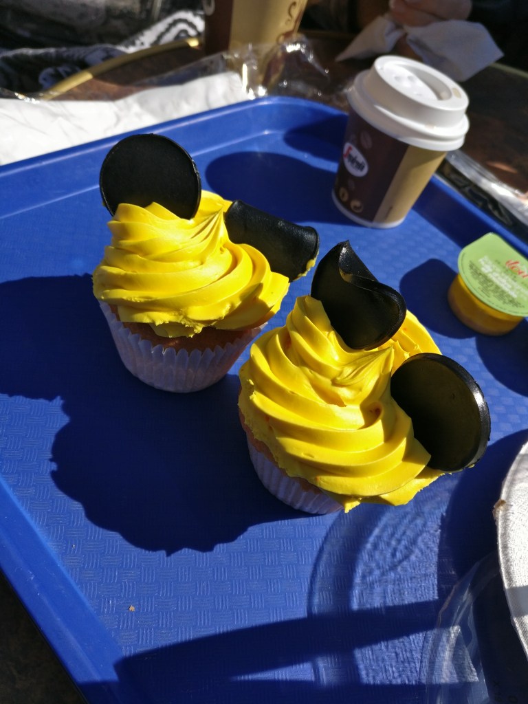 Kaksi keltaisella kuorrutteella päällystettyä kuppikakkua, joihin on laitettu mustat lakritsipyörylät Mikki Hiiren korviksi. Korvat ovat alkaneet löprähtää auringonlämmössä.