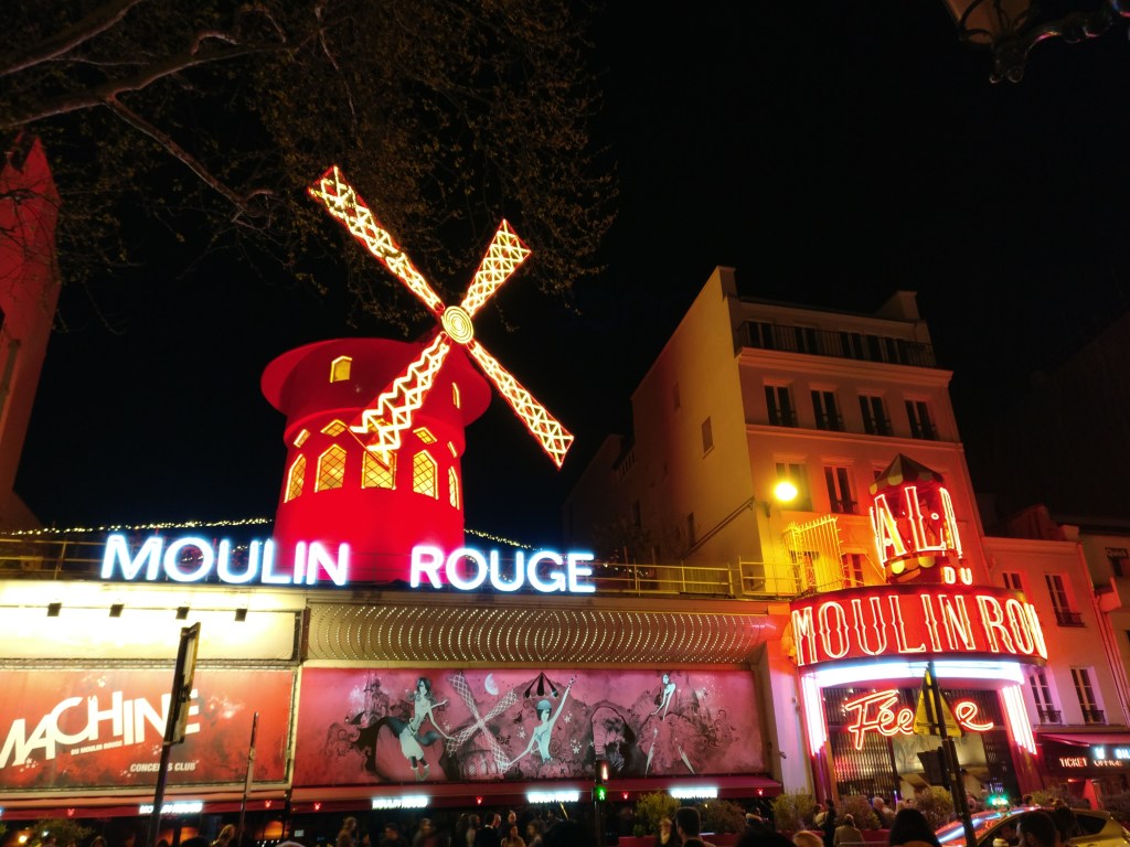 Iltapimeällä kuvattu valokuva, jossa Moulin Rougen punainen mylly hehkuu värikkäiden mainosjulisteiden yllä.