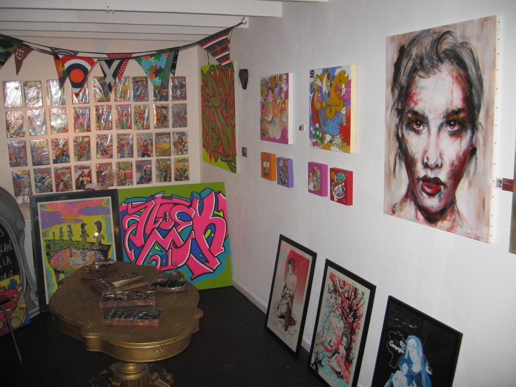 Kuvassa kahvilan nurkkaus, joka on täynnä kuvataidetöitä: osa on ripustettu seinälle, osa nojaa lattialla seinää vasten. Etualalla on pieni pronssisen värinen pöytä.