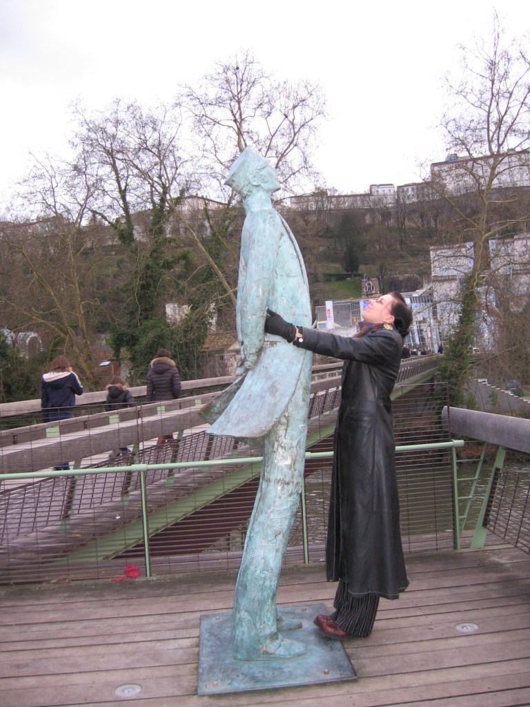 Kuvassa kävelysilta, jonka edessä seisoo sarjakuvahahmo Corto Maltesen patsas. Katja pitelee patsasta vyötäisiltä ja katsoo sitä hymyillen kohti kasvoja (pituusero on huomattava).