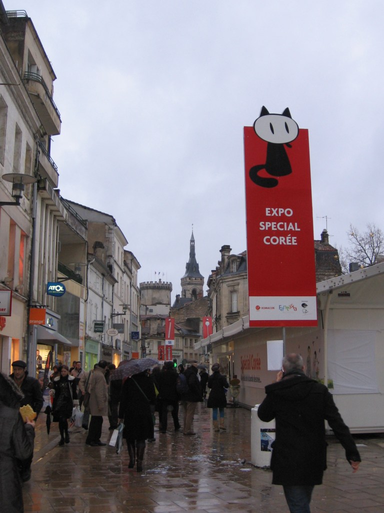 Sateinen kuva kaupungin keskeltä, kävelykadulta, jossa tummiin takkeihin pukeutuneet ihmiset kulkevat. Etuoikealla on iso, punainen pystyjuliste, jossa lukee ranskaksi "Expo special corée" ja sen yläpuolella on festivaalien maskotin, mustavalkoisen piirroskissan kuva.