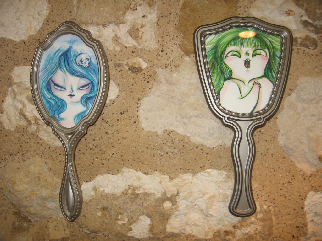 Erikoislähikuva kahdesta käsipeilistä, jotka on ripustettu seinälle. Peiliosan tilalla on piirroskuvat kahdesta naishahmosta: vasemmanpuolimmaisella on sininen tukka ja pääkallo hiuksissa, oikeanpuolimmaisella vihreä tukka ja avoin suu.