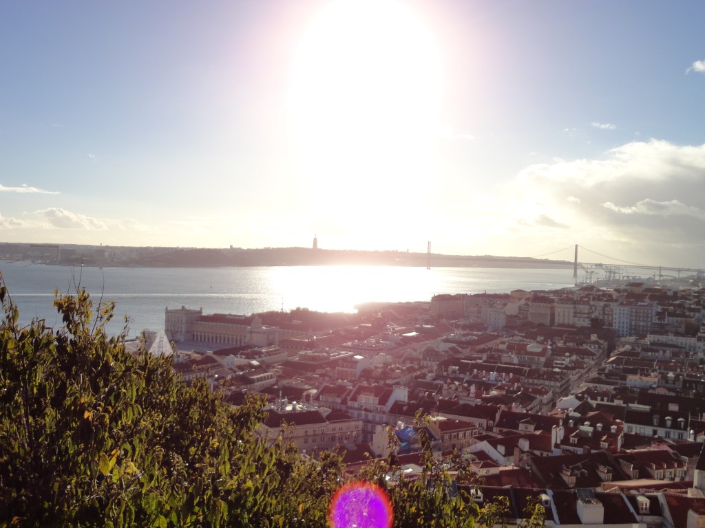 Kuva muurilta kohti kaupunkia ja joenrantaa. Aurinko paistaa suoraan kameraan keskeltä kuvaa, mikä tekee kuvan osin ylivalottuneen näköiseksi.