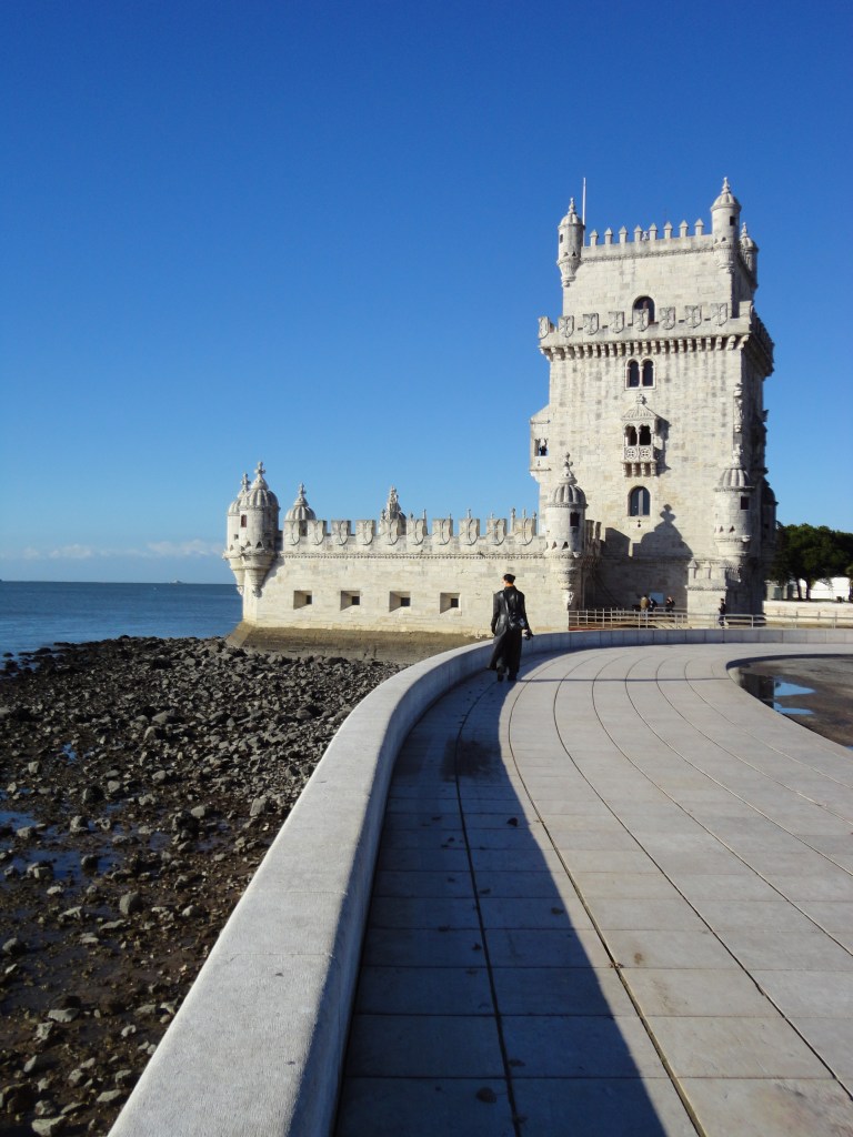 Kuva samasta tornista rantaa myöten otettuna. Kuvassa näkyy vasemmalla kivikkoinen ranta ja itse joki, jonne tornin matalampi "terassimainen" taso ulottuu. Kivetyllä kävelyalueella kävelee Katja mustassa nahkatakissaan kohti tornia.