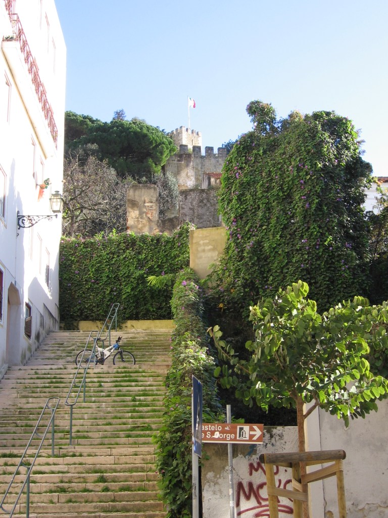 Kuva kapealta kujalta, jolta nousee ruohoa kasvavat rappuset kohti kasvuston peittämää ylätasannetta. Sen takaa pilkottaa kivinen linna, joka on osin puiden peittämä.
Portaiden juurella on kyltti, joka ohjaa Castelo de S. Jorgen suuntaan.
