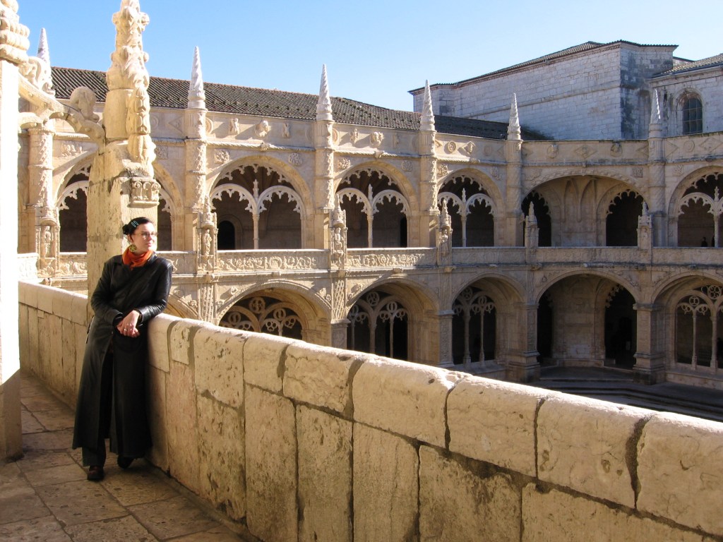 Kuvassa Katja nojailee vaaleaan muuriin luostarin ylätasanteella. Taustalla näkyvät paremmin koristeellisten holvikaarien lisäksi myös kapeat, tornimaiset patsaat, jotka kohoavat katon yli.