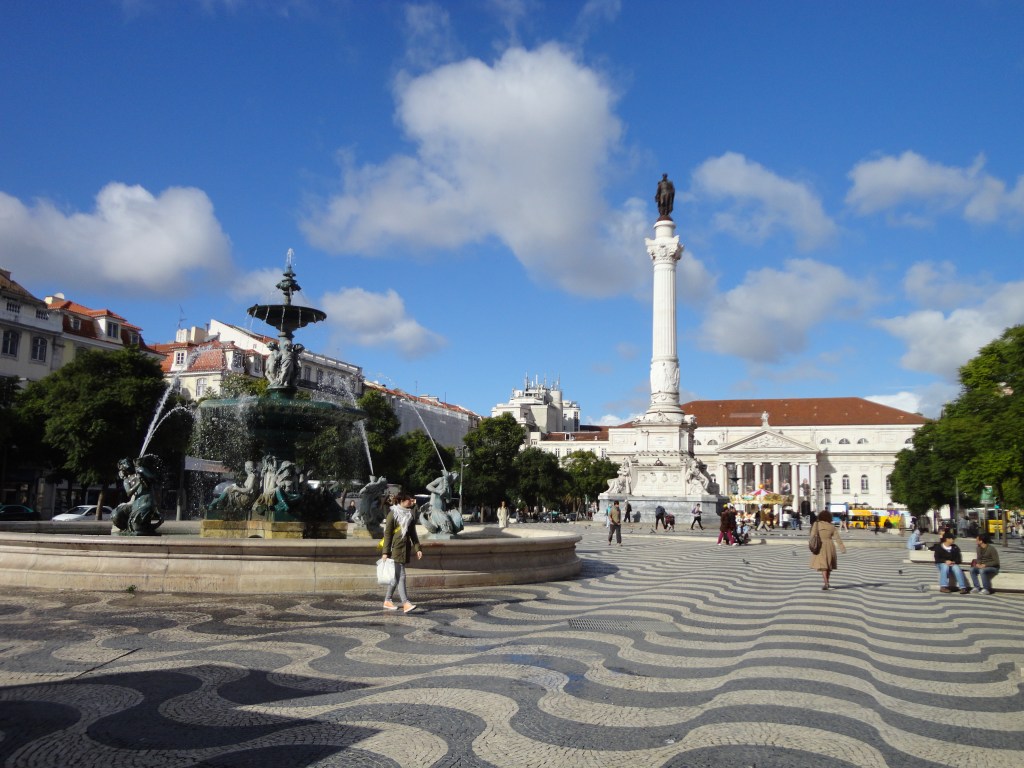 Kuvassa aurinkoinen kaupunginaukio, jonka kiveys on tehty kuvioinnilla aaltoilevan näköiseksi. Etuvasemmalla on iso suihkulähde ja taaempana keskellä kuvaa on valkoinen iso pylväs, jonka päässä seisoo musta hahmo. Takimmaisena on jonkinlainen palatsirakennus, jonka portti on kreikkalaishenkinen eri pylväineen.