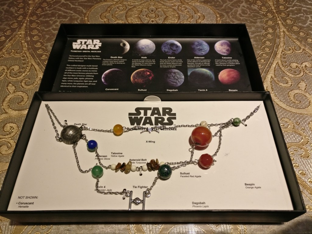 Kuvassa pakkauksessaan oleva kaulakoru, jossa eri korukivet esittävät eri planeettoja Star Wars -universumista. Mukana on myös Kuolemantähti sekä TIE-hävittäjä ja X-siipinen.