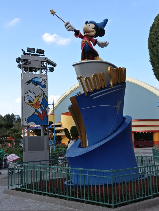 Fantasiasta tuttu velho-Mikki seisoo ison tolpan päällä. Taustalla pylväässä on iso Aku Ankan juliste.