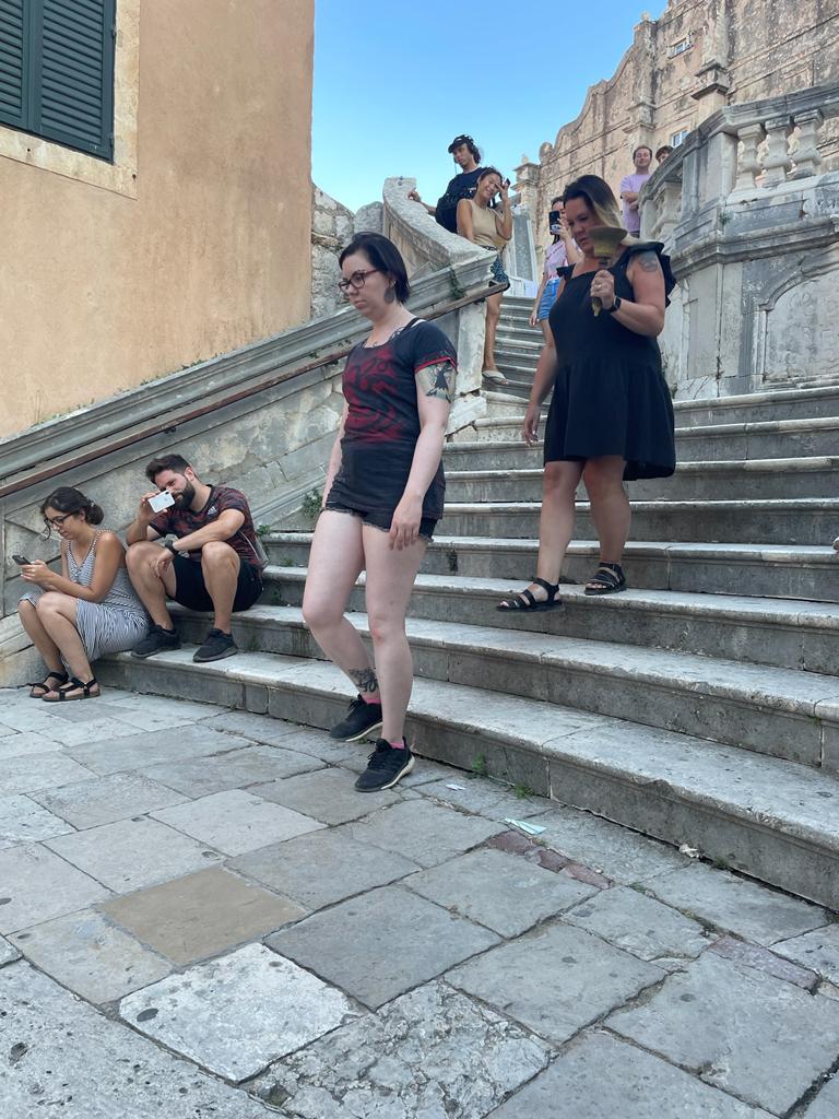 Valokuvassa Katja kävelee vakavana rappusia alas ja hänen takanaan mustamekkoinen nainen pitelee kädessään kilistettävää kelloa.