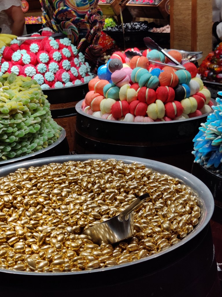 Kuvassa valtavia sammioita täynnä erilaisia karkkeja. Etualalla kultaiseksi värjättyjä munia, taustalla vihreitä sammakoita, jättimäisiä macaron-leivoskarkkeja, isoja mansikoita ja valtavia tikkareita.