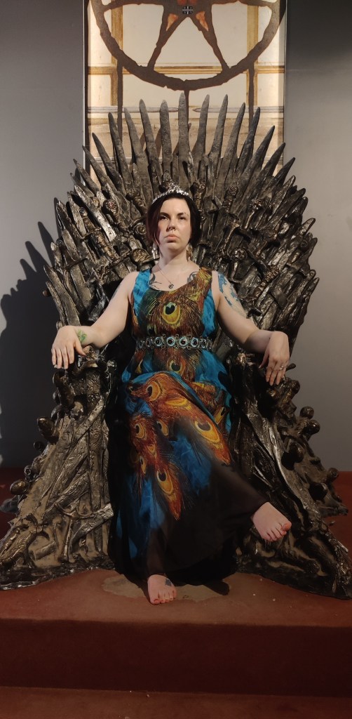 Kuvassa Katja rautavaltaistuimella. Katjalla on tuima ilme, kruunu päässä ja yllä pitkä, riikinkukonsulkakuvioinen mekko.