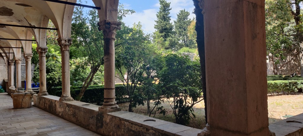 Kuvassa sisäpiha, jossa paljon puita, mutta karun näköinen nurmikko. Kuva on otettu katetulta, kivetyltä kävelyreitiltä, jonka kattoa kannattelee useampi, koristeellinen pylväs.