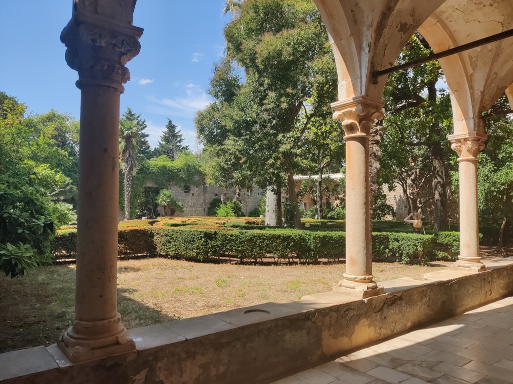 Puutarhan kuva hieman eri kulmasta: tästä näkee tasoitetut pensasaidat, erilaiset puut, kuten palmut, ja lähietäisyydeltä koristeelliset pylväät. Aurinko paistaa, ruohikko näyttää paahtuneelta.