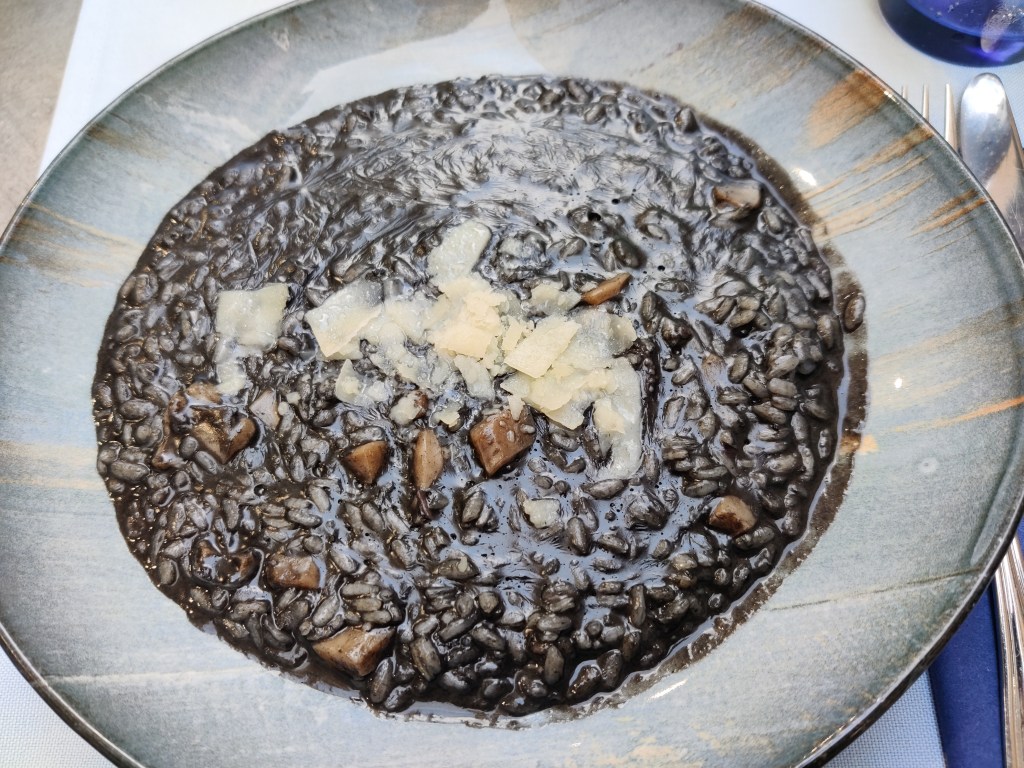 Kuvassa lautasella oleva risottoannos, joka näyttää väriltään ruskeanmustalta. Risotto on kosteaa ja siinä on vaaleanruskeita mustekalanpaloja. Sen päällä on vaaleita juustohiutaleita.