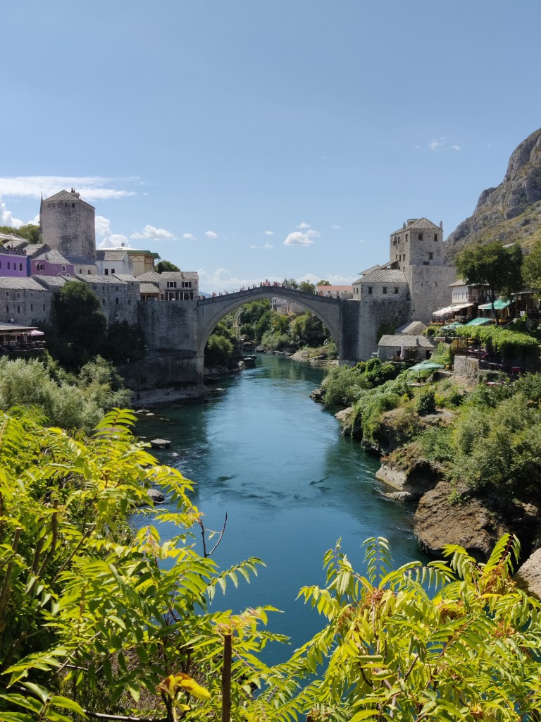 Valokuvassa pyöreä silta sinisen, kapean joen yllä. Etualalla paljon vaaleanvihreitä pensaita. Kivisillan kummallakin puolella on useita linnoitusmaiseia rakennuksia. Joen rannalla on vihreitä pensaita. Oikealla maisema kohoaa korkeaksi, karuksi kukkulaksi. Taivas on lähes pilvetön. Sillalla näkyy heikosta ihmisiä.