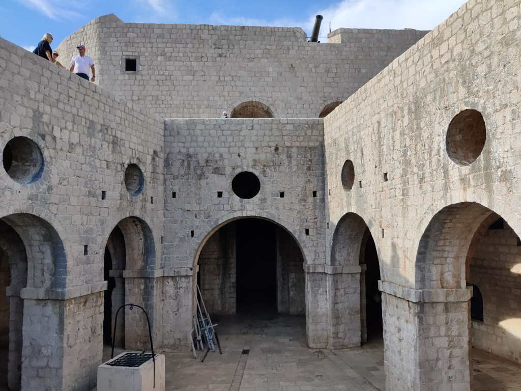 Kuvassa kivisen linnoituksen sisäpiha, jossa useita kaarioviaukkoja, joiden päällä pyöreät ikkuna-aukot. Linnoituksella on ylätasanne, jossa käyskentelee pari ihmistä.