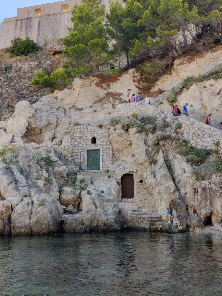 Kuvassa rantakallio, jossa näyttää olevan kaksi eri tasoilla olevaa ovea. Rinteestä erottuu kaksi eri porrastasoa, joilla kulkee ihmisiä.
Kallion päällä olevasta linnoituksesta näkyy pieni osa.