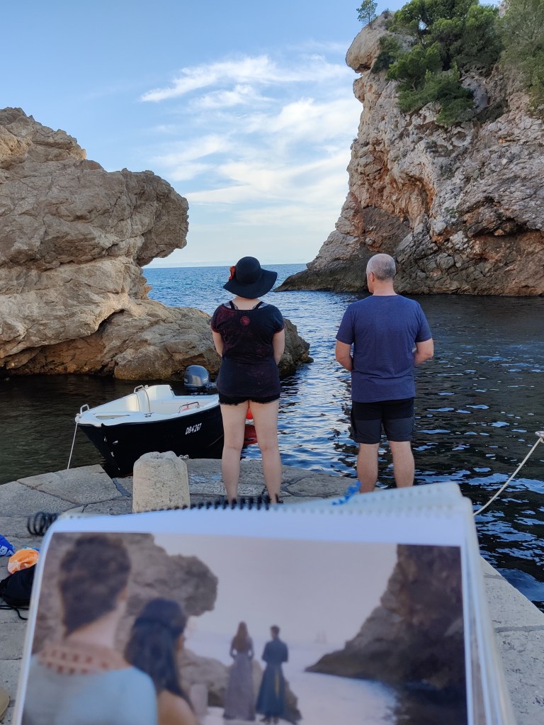 Kuvassa mies ja nainen seisovat selin kameraan kivisellä laiturilla. Kummassakin reunassa isot rantakalliot rajaavat näkyvyyttä. Kuvan etualalla on lehtiö, jossa on valokuva Game of Thrones -tv-sarjasta. Kuvassa seisovat ihmiset matkivat valokuvan henkilöiden asentoja.