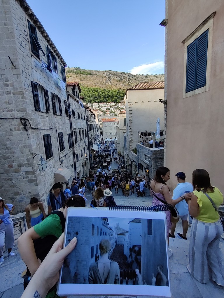 Kuva ison portaikon yläpäästä. Portaat jatkuvat alaspäin kapeaa katua pitkin, kummallakin puolella on korkeita rakennuksia. Portaat ja katu ovat täynnä ihmisiä. Kuvan etualalla käsi pitelee lehtiötä, jossa on kuvattu Game of Thronesin Cersei seisomassa tällä samalla paikalla.