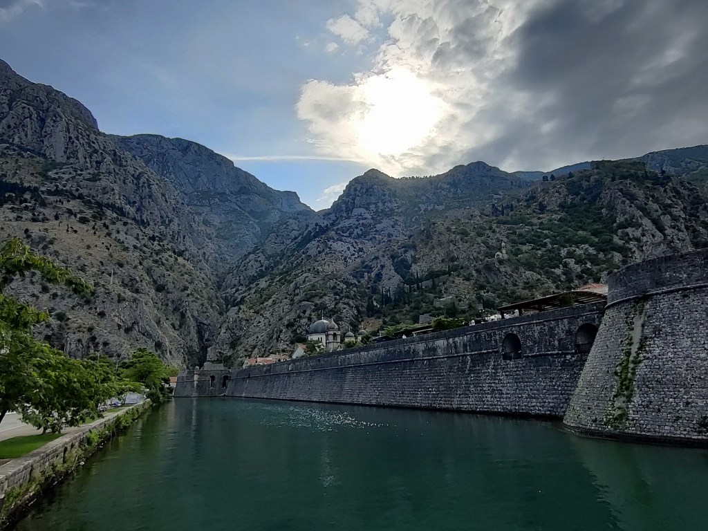 Kuva vallihaudasta hieman eri kohtaa. Vallihaudan toisella reunalla näkyy puiden reunustama kävelytie. Muuri päättyy vuorten juurelle. Muurin sisäpuolella erottuu kirkontornin kupoli. Korkealla vuorenrinteessä voi nähdä vaalean kirkon.
