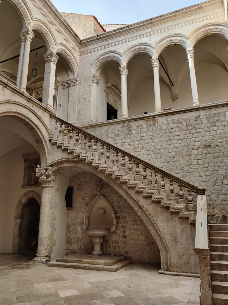 Kuvassa kivisen palatsin sisäpihan portaikko. Portaiden alla on pieni vesiallas. Portaat johtavat ylätasanteelle, jonka kattoa kannattelee lukuisa määrä kapeita pylväitä. Värimaailma on vaaleanruskea. Palatsin tässä osassa ei ole kattoa, vaan kuvan yläreunassa näkyy taivas.