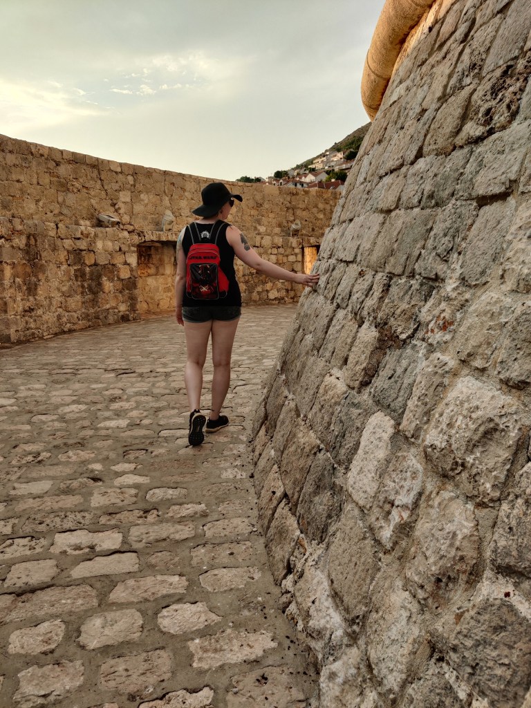 Kuvassa Katja kulkee selin kameraan kiveyksellä. Taustalla on korkea muuri. Katjan oikea käsi hyväilee oikeassa reunassa ylöspäin kohoavaa tornia, joka jo kuvassa kaartuu vahvasti oikealle.