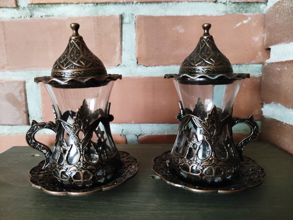 Kaksi perinteistä lasista teekuppia, joiden alaosaa kehystää koristeellinen, metallinen "tukiranka", jossa on kahva. Kupit seisovat koristeellisilla aseteilla ja kummankin päällä on pieni lautanen, jonka päällä on puolestaan pieni suojakupu.