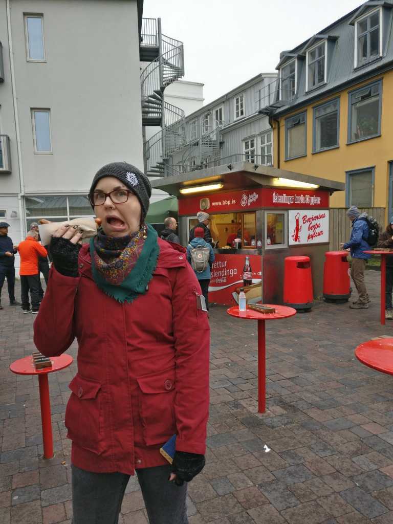 Kuvassa Katja, joka on haukkaamassa suu vinossa paperiin käärittyä hodaria. Taustalla on punaisia seisomapöytiä ja punainen hodarikioski, jonne on jo melkoinen jono.