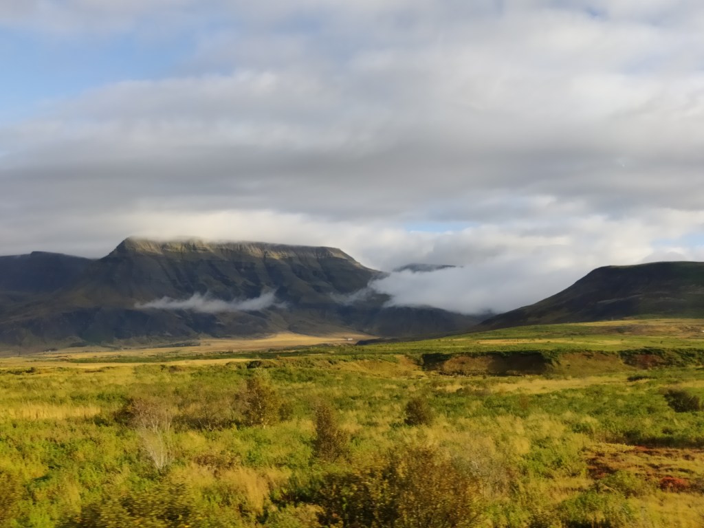 Maisemakuva, jossa etualalla matalaa pensaikkoa ja taustalla kohoavaa vuoristoa. Valkoiset pilvet ovat niin matalalla, että ne kulkevat vuorten edustalla.