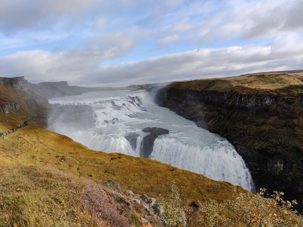 Kahden kellertävän vihreän kallion keskellä valtava vesiputous, jossa vesi putoaa kallioiden väliin näkymättömiin. Vasemmassa reunassa näkyy jono ihmisiä kulkemassa polkua pitkin kohti vesiputouksen huippua.