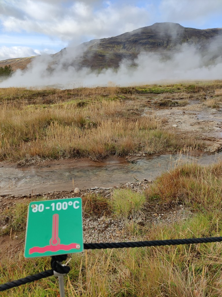 Kuvassa köydellä eristettyä kostea, ruohikkoinen alue, joka höyryää. Tolpassa on vihreä kyltti, jossa lämpömittarin kuva ja teksti 80 - 100 Celcius-astetta.